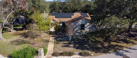 A house with a stone facade, a solar panel roof, and a well-maintained garden with a small tree and shrubs.