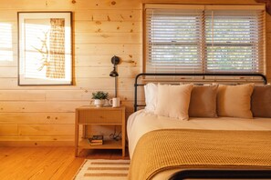 A wooden-paneled room with a bed, a bedside table with a plant and books, and a window with blinds. - Whippoorwill Estate - Spa | Mini Golf | B Ball (Mineral Bluff)