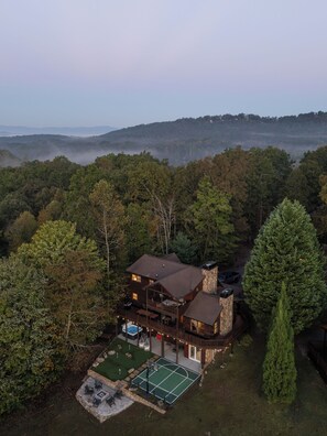 A two-story wooden house with a stone chimney, surrounded by dense forest and a tennis court. - Whippoorwill Estate - Spa | Mini Golf | B Ball (Mineral Bluff)