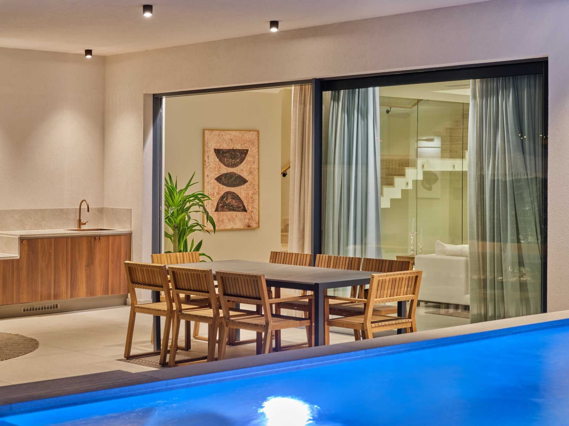 Un área de piscina interior moderna con una mesa y sillas de madera, un lavamanos y un amplio ventanal con cortinas.
