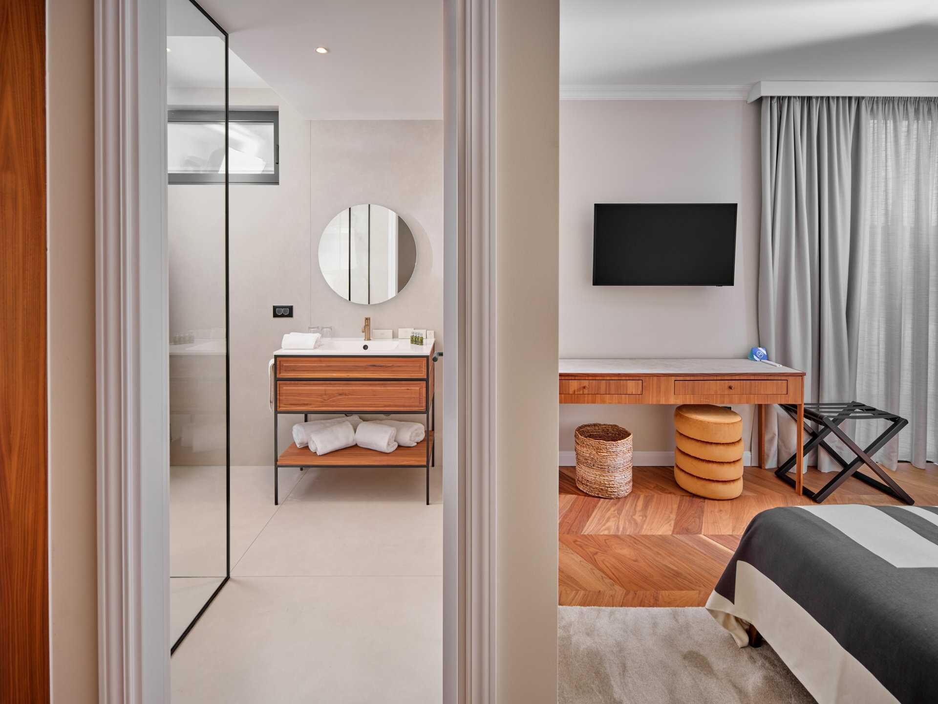 A modern hotel room with a wooden vanity, a round mirror, a flat-screen TV, a desk, and a bed with a striped bedspread.