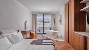 A modern hotel room with a large bed, a vanity with a mirror, a bathtub, and a view of a coastal town. - Luxury Villa Lunea with Pool VII (Trogir)