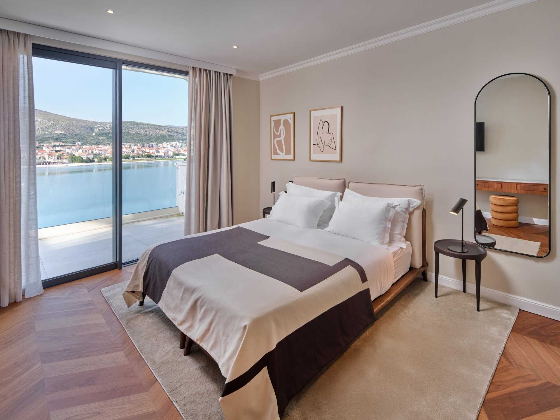A modern bedroom with a large bed, a view of a body of water, and a cityscape.