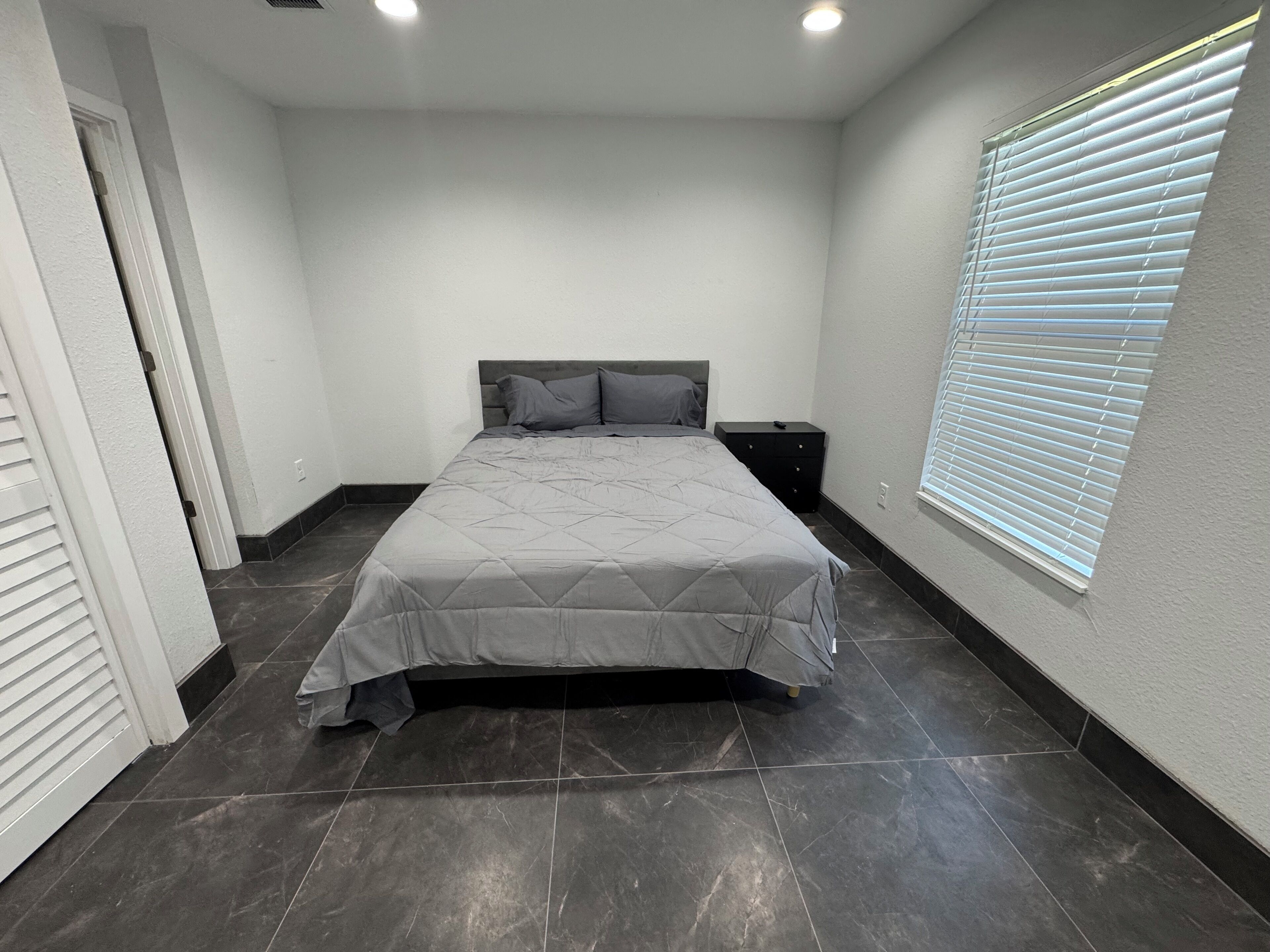 A bedroom with a bed, a nightstand, a window with blinds, and a door.