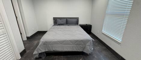 A bedroom with a bed, a nightstand, a window with blinds, and a door.