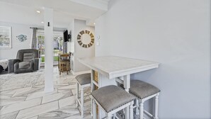 En rummelig opholdsstue med et hvidt spisebord og bænk, en grå lænestol og et fjernsyn monteret på væggen.