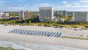 Une plage bordée de rangées de parasols bleus et jaunes, un grand bâtiment en arrière-plan et une vue sur l’océan.