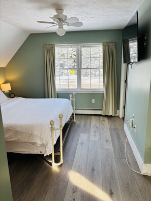 Un dormitorio con cama, ventilador de techo, ventana con cortinas y televisor en la pared.