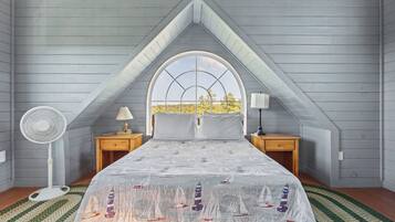 Ein Schlafzimmer mit schrägen Wänden, einem großen Fenster, einem Bett mit einem marineblauen Bettbezug, zwei Nachttischen mit Lampen und einem Stehlampenventilator.