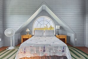 A bedroom with a sloped ceiling, a large window, a bed with a nautical-themed bedspread, two bedside tables with lamps, and a standing fan. - Coastal Lighthouse Retreat 4mi to Acadia
Sleeps 16 guest (Tremont)