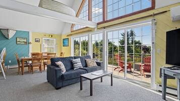 A spacious living room with a blue sofa, a dining area with wooden chairs, and a large sliding glass door leading to an outdoor patio with red chairs.