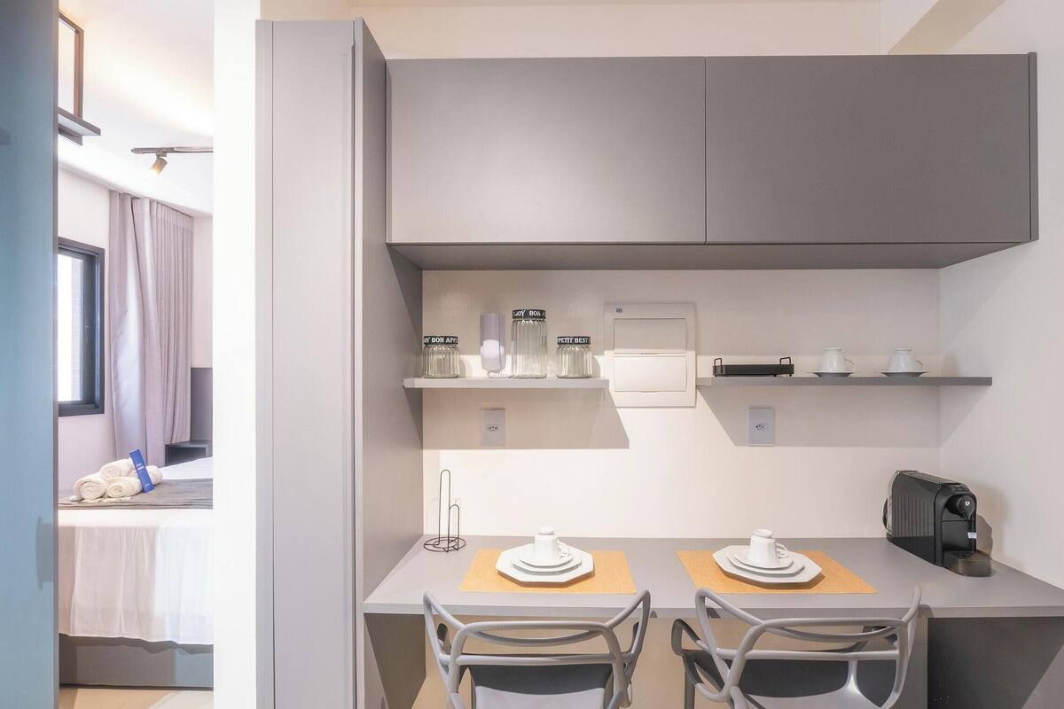 A compact kitchenette with a coffee machine, a small table with chairs, and a window with curtains.