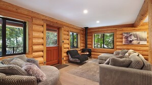 A log cabin interior with a fireplace, multiple seating areas, and large windows offering views of the forest. - Wintergreen Lodge, Mount Washington - Hot Tub (Comox-Strathcona C)