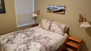 Een slaapkamer met een bed, een nachtkastje, een lamp, een schilderij aan de muur en een raam met jaloezieën.