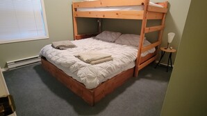 Een stapelbed met houten frame, wit beddengoed en een klein nachtkastje met een lamp.
