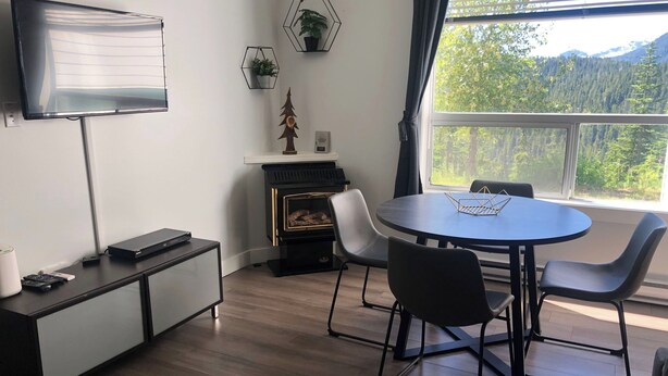Quarto moderno com uma mesa redonda, cadeiras, TV de tela plana, lareira e janela com vista para a floresta.