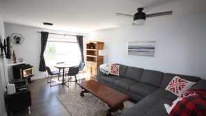 Uma sala de estar moderna com um sofá cinzento, uma mesa de centro de madeira, uma mesa de jantar com cadeiras, um ventilador de teto e uma janela com cortinas.