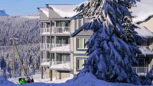 A multi-story hotel with balconies, surrounded by snow-covered trees and a mountainous backdrop. - Strathcona Views - Blueberry (Comox-Strathcona C)