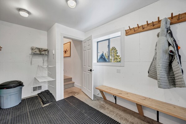 Une pièce bien éclairée, avec un banc en bois, un porte-manteau, une porte donnant sur un escalier et une fenêtre offrant une vue sur les arbres.