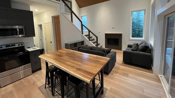 Una cocina moderna con una mesa de madera y taburetes altos, una escalera que sube a un nivel superior, y una sala de estar con chimenea y sofá.