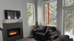 Una sala de estar moderna con chimenea, sofás grises y ventanas amplias que dan a un paisaje de árboles.