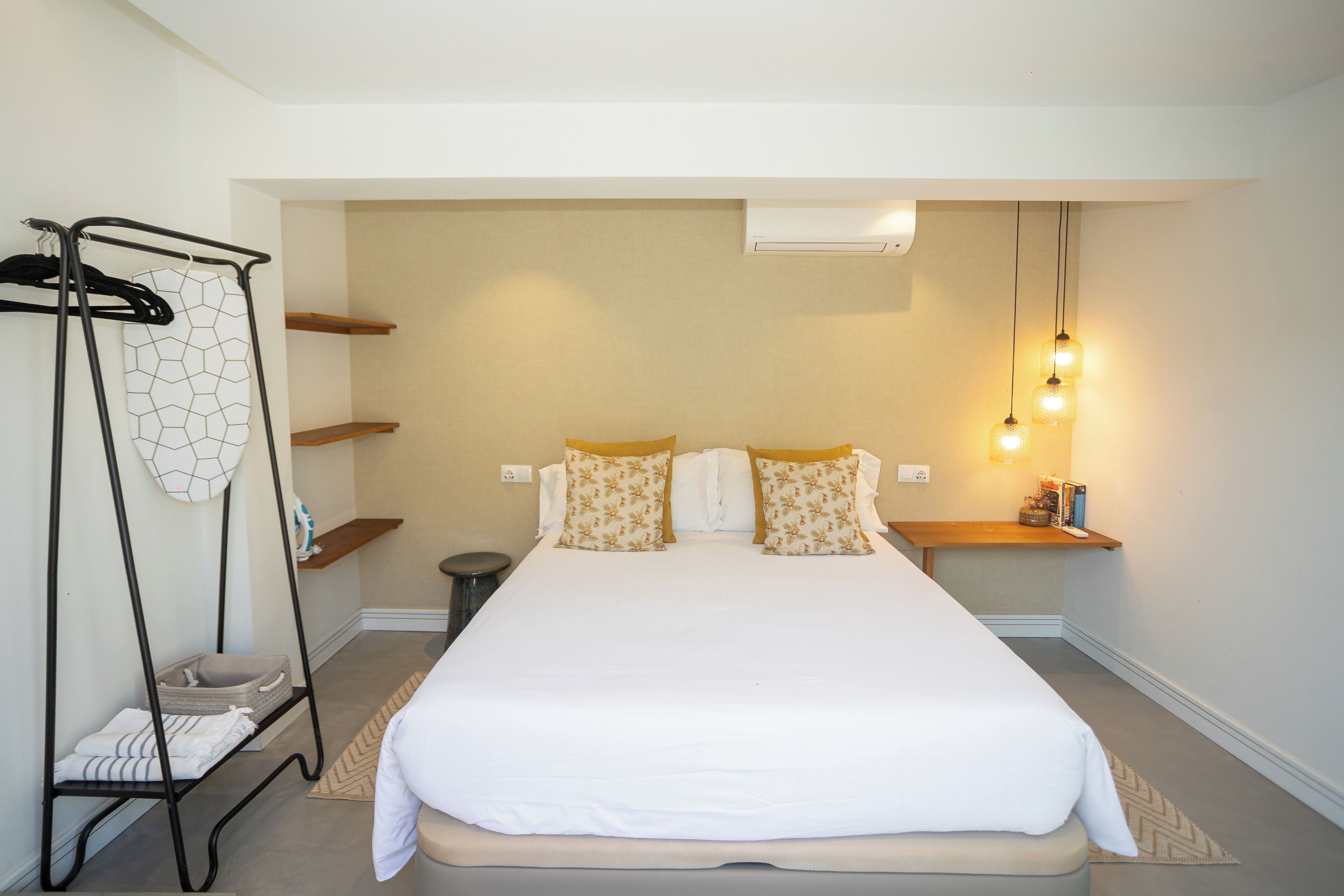 Een slaapkamer met een bed, een ophangrek voor wasgoed, een klein plankje en een aan de muur bevestigde airconditioner.