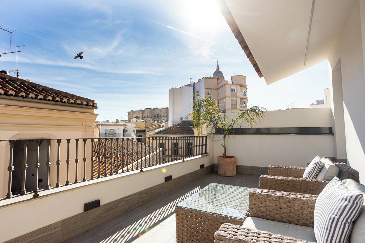A rooftop terrace with wicker furniture, a potted plant, and a view of surrounding buildings.