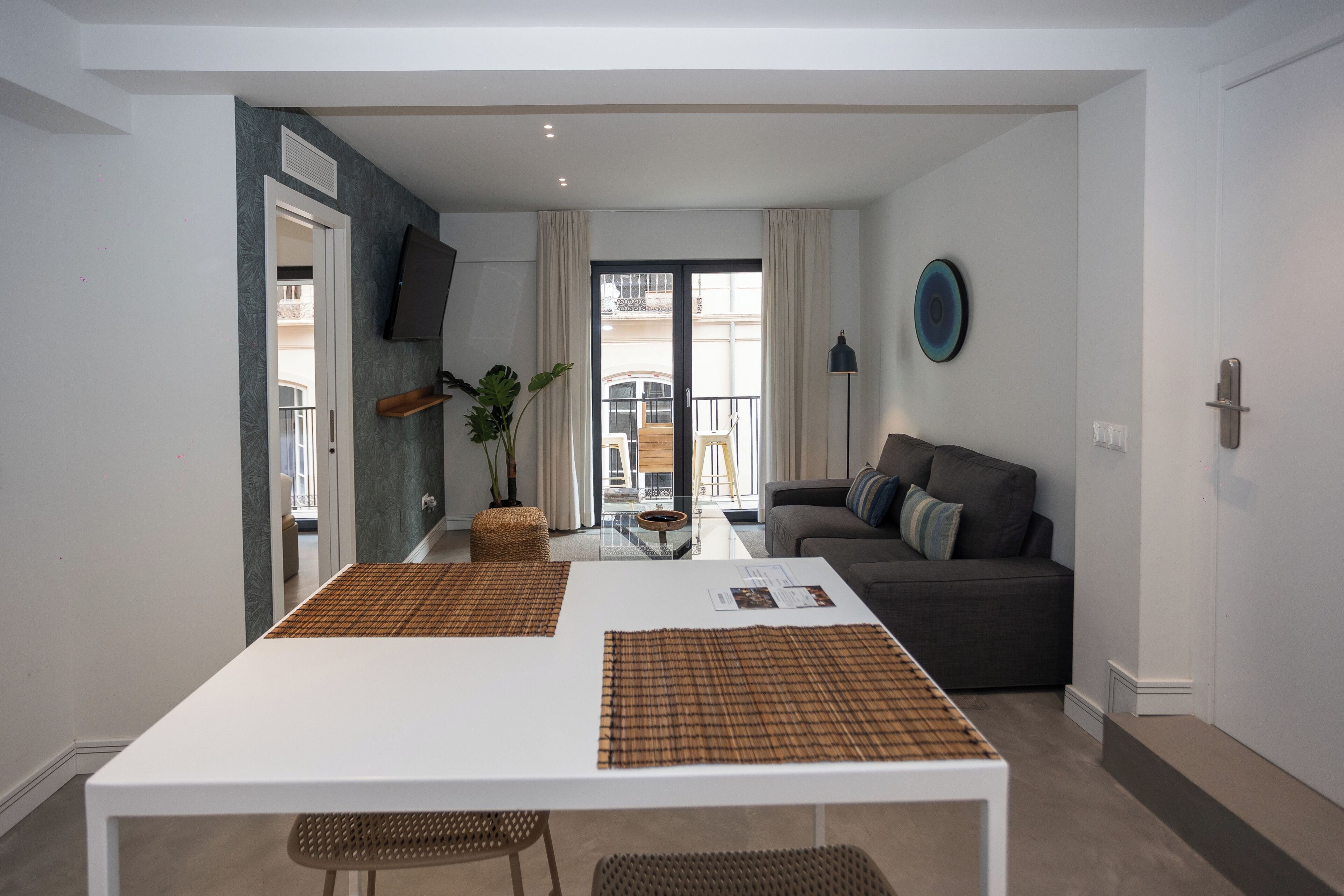 A modern living room with a sofa, a dining table with placemats, a TV mounted on the wall, and a balcony with a view.