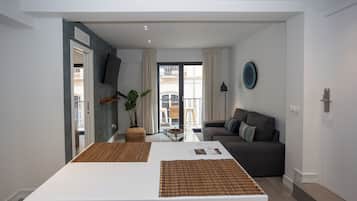 A modern living room with a sofa, a dining table with placemats, a TV mounted on the wall, and a balcony with a view.