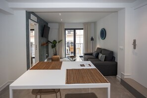 Un salon moderne avec un canapé, une table à manger agrémentée de sets de table, un téléviseur fixé au mur et un balcon offrant une vue agréable.