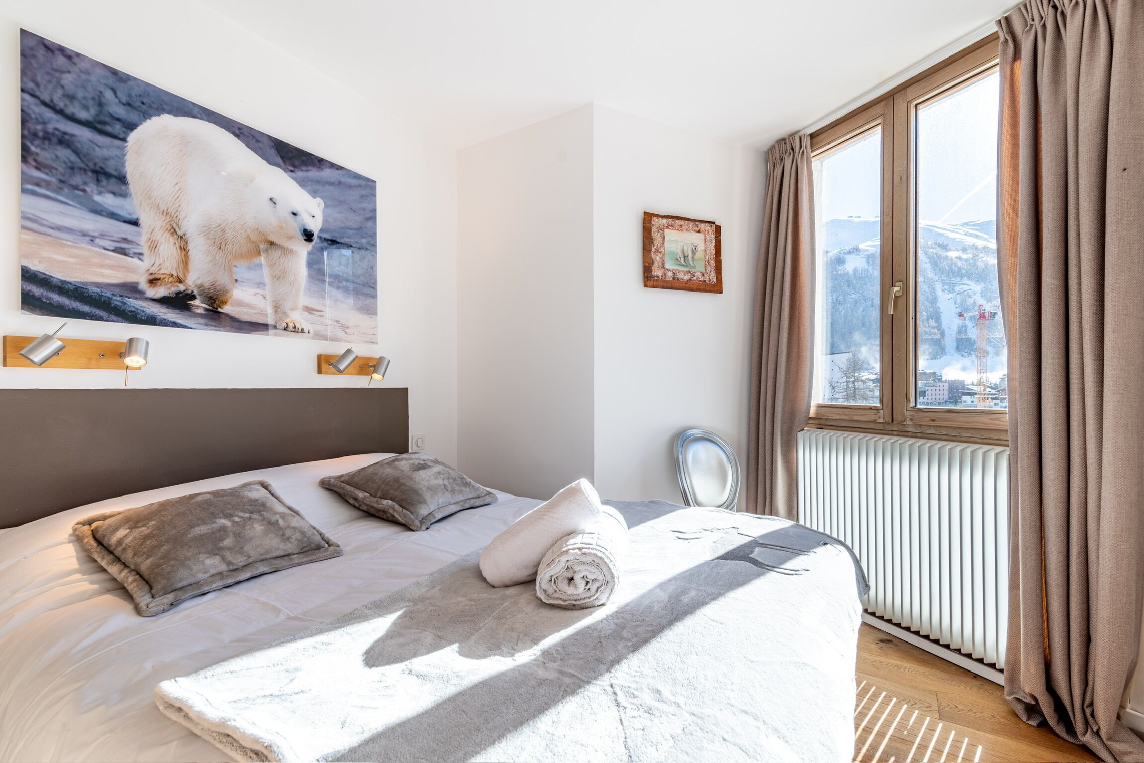 Une chambre avec une grande fenêtre, un lit sur lequel est posée une serviette roulée, un tableau représentant un ours polaire et un radiateur.