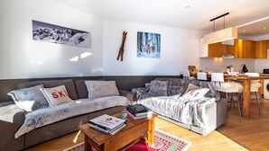 A modern living room with a sofa, a coffee table with books, and a kitchen area with wooden cabinets and a dining table. - NEW - Family apartment for 5-6 people with exceptional view of Val d'Isère (Val-d'Isère)