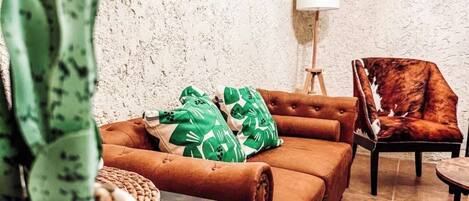 A cozy living room with a brown leather sofa, green patterned pillows, a potted plant, and a framed artwork on the wall.