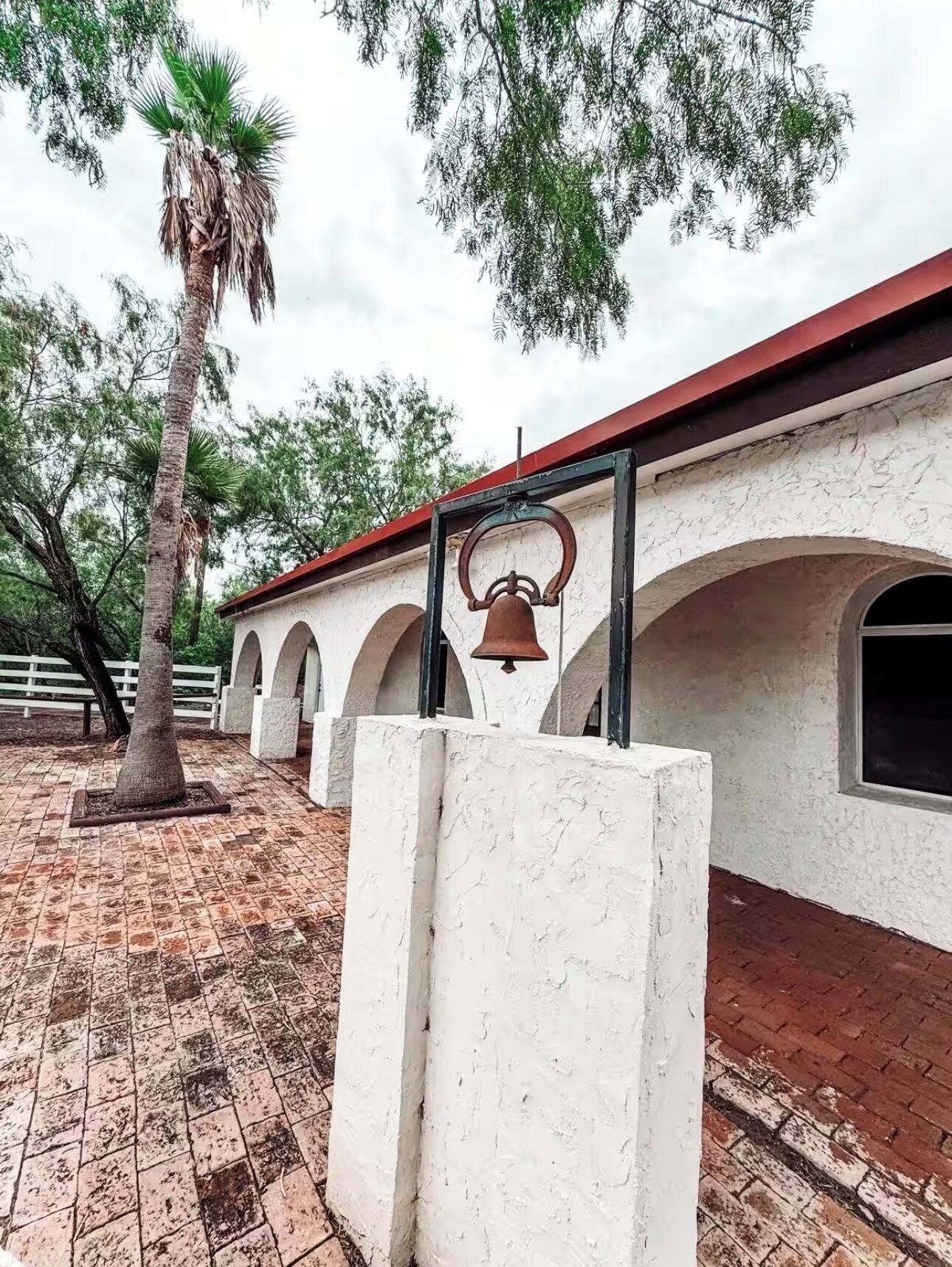 Um edifício com telhado vermelho, paredes brancas e janelas em arco. Há uma palmeira e um sino em primeiro plano.