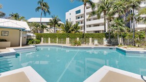 A swimming pool with a tiled edge, surrounded by palm trees and a white building with balconies. - Ocean view 1BR, 1 min to beach, shared pool (Rum Point)