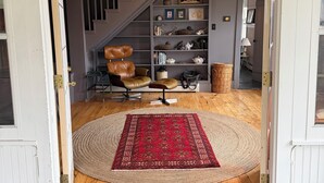 Un vestíbulo con suelo de madera, un tapete rojo estampado, un sillón de cuero y una estantería con varios objetos decorativos.