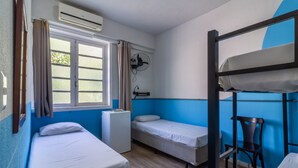 Quarto de dormitório com beliches, escrivaninha, cadeira, janela com cortinas e ventoinha fixada na parede.
