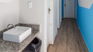 Corredor estreito com banheiro à esquerda, com pia e lixeira. As paredes são pintadas de branco e azul, e há uma porta com um sinal de número.