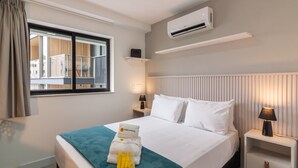 Ein Hotelzimmer mit einem Bett, einem Fenster mit Vorhängen, einer Klimaanlage, einem Nachttisch mit Lampe und einem an der Wand befestigten Regal.