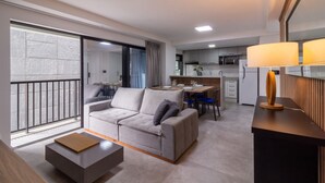 Ein modernes Wohnzimmer mit einer Couch, einem Couchtisch und einem Essbereich mit Stühlen und Tisch. Im Hintergrund ist eine Küche mit Schränken und Geräten zu sehen.