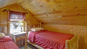 Une petite pièce chaleureuse aux murs en bois et au plafond en pente. On y trouve un lit simple avec une couverture rouge, une petite table avec une lampe et une fenêtre avec un rideau.