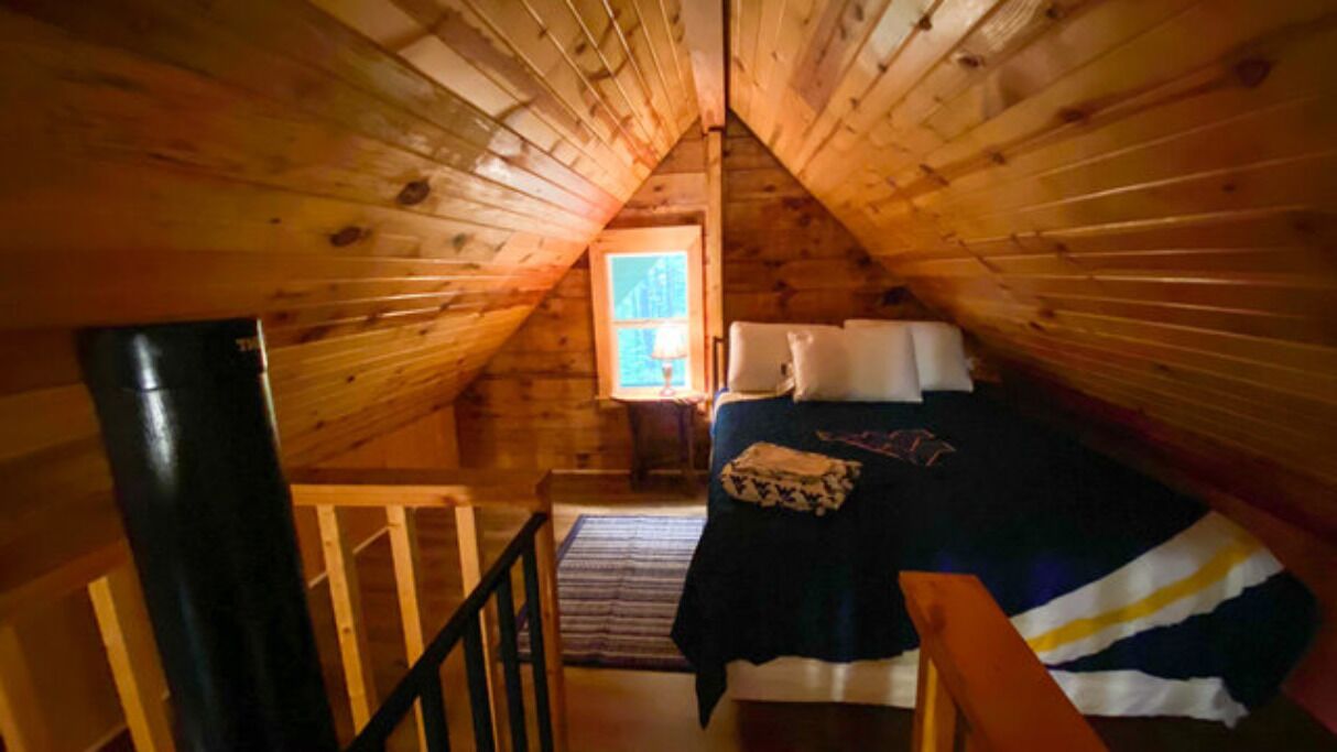 A cozy attic bedroom with a sloped ceiling, a bed with pillows, a nightstand, and a window letting in natural light.