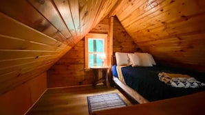 Une chambre mansardée chaleureuse, avec un plafond en pente en bois, un lit simple avec des oreillers, une table de chevet et une fenêtre laissant entrer la lumière naturelle.