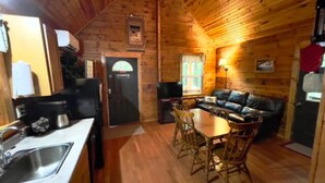 L’intérieur chaleureux d’un chalet, avec un revêtement mural en bois, un coin repas avec des chaises, un canapé et une cuisine équipée d’un évier et d’un réfrigérateur.