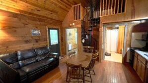 L'intérieur d'un chalet en bois est aménagé avec un canapé en cuir noir, une table à manger en bois entourée de chaises et un escalier menant à une mezzanine.