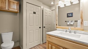 A bathroom with two white doors, a toilet, a sink with a marble countertop, and a mirror with a light fixture. - Brand New Cozy Glacier Cabin (Coram)