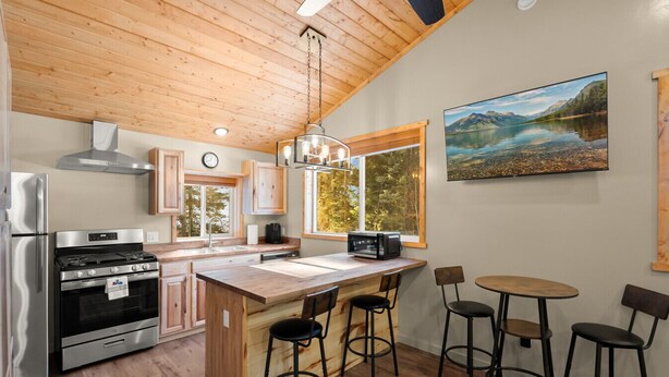 A modern kitchen with wooden cabinetry, a stainless steel refrigerator, an oven, and a microwave. There is a wooden island with bar stools and a dining table. Large windows provide natural light and a view of the outdoors. - Brand New Cozy Glacier Cabin (Coram)