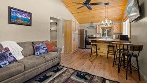 A cozy living room with a sofa, a coffee table, and a patterned rug. A kitchen with wooden cabinets and a bar area is visible in the background. - Brand New Cozy Glacier Cabin (Coram)