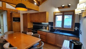 Una cocina con armarios de madera, una mesa de comedor con sillas, una estufa negra y una ventana con cortinas.