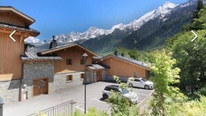Una cabaña de madera con paredes de piedra, un estacionamiento y montañas al fondo.